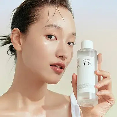 Woman with dewy skin holding clear 77% toner bottle, Korean skincare product