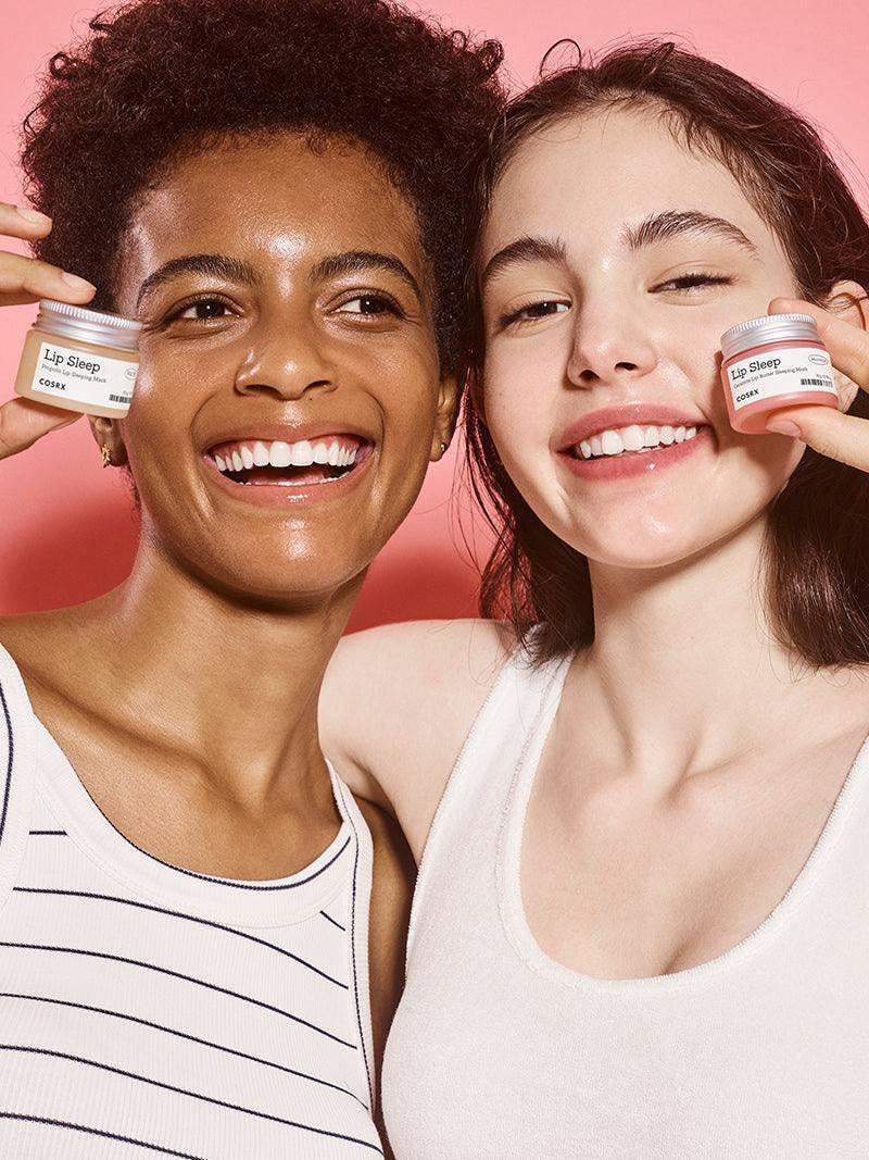 Two smiling women holding COSRX Lip Sleep cream against pink background, skincare concept