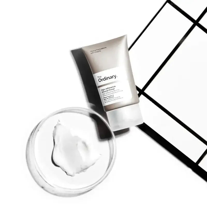 The Ordinary High-Adherence Silicone Primer tube next to a clear dish with creamy skincare product