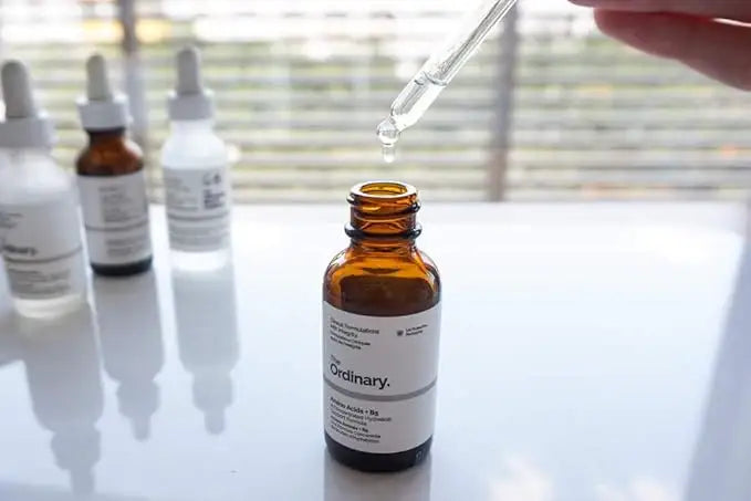 Dropper dispensing serum into brown glass skincare bottle with other serums in background