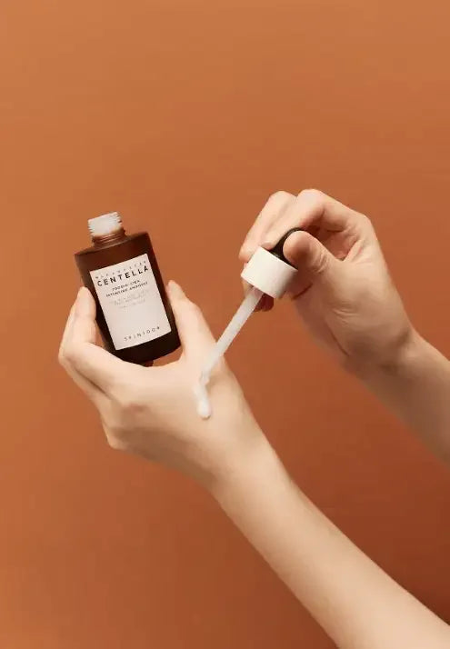 Hand applying Centella skincare serum with dropper against a brown background