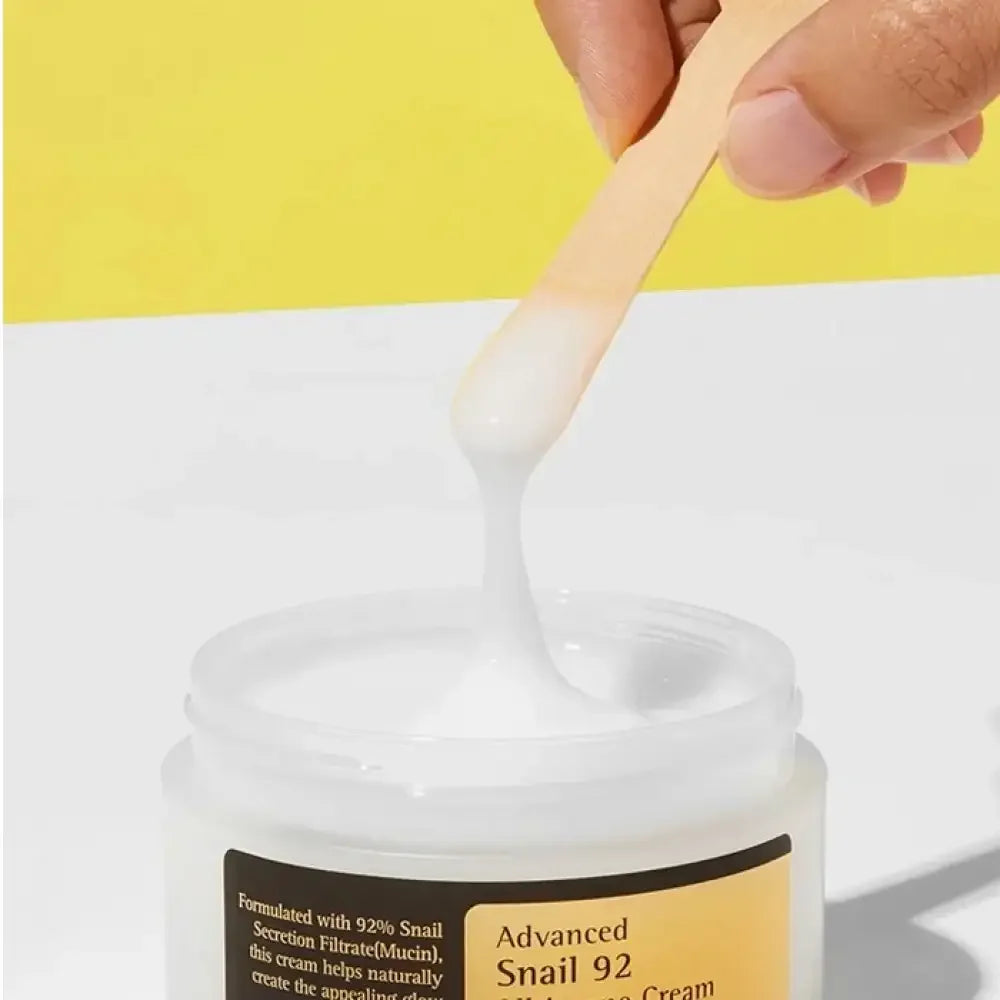 Hand holding spatula with white snail mucin cream above open jar on white and yellow background