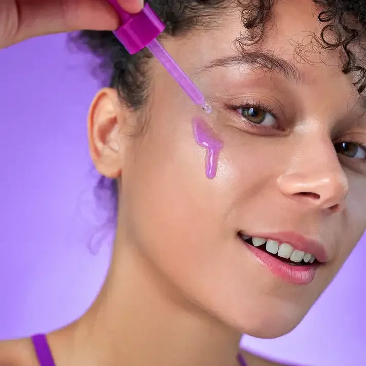 Woman applying purple skincare serum to cheek with dropper, glowing skin, purple background