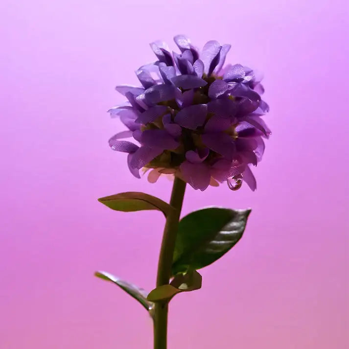 Purple artificial flower with green leaves against a pink gradient background